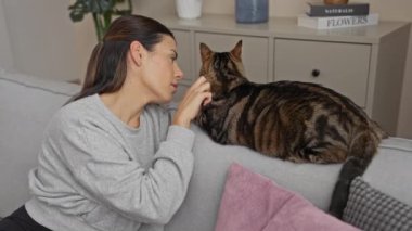 Genç bir İspanyol kadın sıcak bir oturma odasında kanepede kedi yavrusuna sarılıp öpüyor. Kapalı bir ortamda sevgi ve rahatlık sergiliyor..