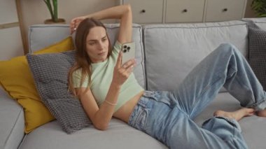 Sarı ve gri yastıklı bir kadın evinin oturma odasındaki telefona bakarken kanepede esniyor..