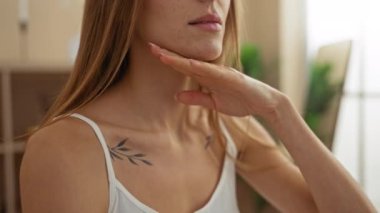 Sarışın ve dövmeli güzel bir genç kadın bir sağlık salonunda oturuyor, nazikçe yüzüne dokunuyor, sakin ve kapalı bir spa havası yayıyor..