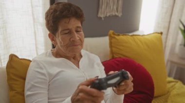 Kısa saçlı, beyaz tişörtlü, olgun bir İspanyol kadın yatak odasında, bir kumandayla video oyunu oynamaya odaklanmış..