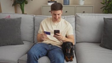 Evde oturan İspanyol bir adam, elinde kredi kartı ve telefon, bir kanepede oturuyor. Tekel, dachshund köpeği oturma odasında, modern dekorlu bir dairede oturuyor..