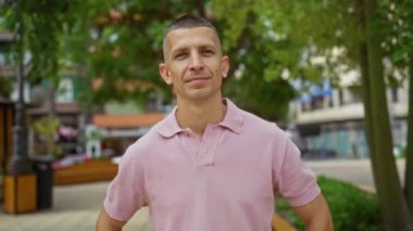 Pembe polo tişörtlü genç adam şehir parkının dışında duruyor. Şehir arka planında kendine güvenen bir ifade var.