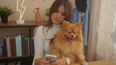 Gülümseyen bir kadın, rahat bir oturma odasında akıllı telefon kullanırken Pomeranian köpeğiyle vakit geçirmekten zevk alır..