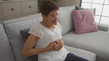 Oturma odasında gri bir koltukta oturan, gözlük takan ve midesini evde tutan, kısa saçlı yaşlı İspanyol kadın..