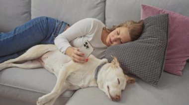 Bir kadın, rahat bir evde, elinde akıllı telefonuyla uyuyan köpeği ile kanepede dinlenir..