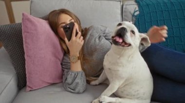 Genç bir kadın köpeğiyle kanepede dinleniyor, rahat bir oturma odasında telefonuna sesli mesaj yolluyor..