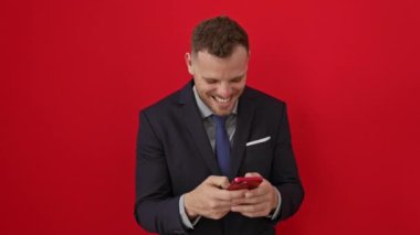 Takım elbiseli genç bir İspanyol, telefonunda komik bir şakayla gülerken yüksek sesle gülüyor, izole edilmiş kırmızı bir arka plana gülümsüyor.