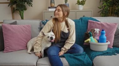 Sarışın, beyaz bir kadın evcil köpeğiyle oturma odasındaki kanepeye oturur. Etrafı temizlik malzemeleri ve yastıklarla çevrilidir..