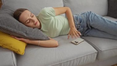 Modern bir oturma odasındaki kanepede uyuyan genç bir kadın elinde akıllı bir telefonla rahat bir dairenin huzurlu atmosferini yakalıyor..