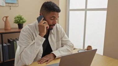 Genç adam, modern bir iç mekanda telefonla konuşurken evde dizüstü bilgisayarla çalışıyor..