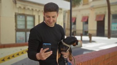 Dachshund köpeği tutan genç bir adam güneşli bir şehir sokağında selfie çekiyor..
