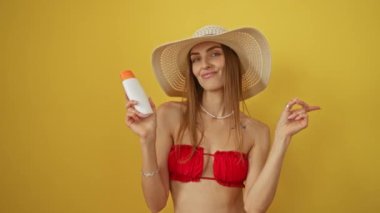 Bikinili kadın güneş kremi tutuyor, gülümsüyor ve el ve parmağıyla yüzünü gösteriyor, izole edilmiş sarı arka planda, mutlu beyaz kadın neşeli bir yaz havası sergiliyor.