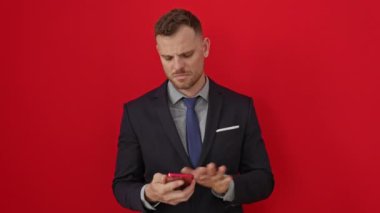 Kuşku ve kafa karışıklığını ifade eden takım elbiseli, cahil İspanyol genç adam izole edilmiş kırmızı arka plan üzerinde akıllı telefon kullanıyor.
