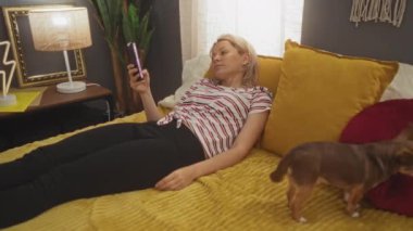Sarışın genç bir kadın, sıcak bir yatak odasındaki bir yatağa uzanıyor, elinde bir telefon, yanında da Chihuahua köpeği duruyor..