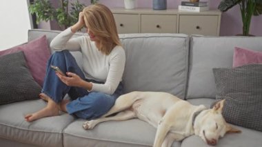 Akıllı telefonlu endişeli yetişkin kadın oturma odasındaki kanepede uyuyan köpeğin yanında oturuyor.