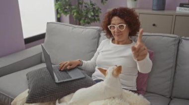 Orta yaşlı bir kadın oturma odasında kanepede oturuyor, kucağında bir chihuahua köpeği, dizüstü bilgisayar kullanıyor, ve parmağını kaldırıyor..