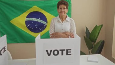 Yaşlı bir İspanyol kadın, arka planında Brezilya bayrağı olan ve seçim odasında oy verme kabinlerinin yanında duruyor..