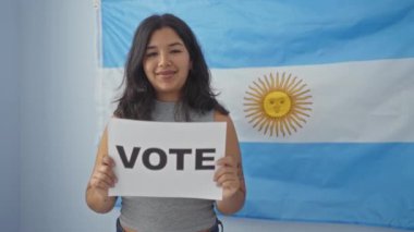 Genç bir İspanyol kadın, arka planda Arjantin bayrağı olan kapalı bir alanda gülümseyerek seçim sürecine katılmanın önemini sembolize eden bir oy pusulası tutuyor..