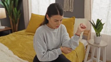 Genç bir İspanyol kadın rahat bir yatak odasında bir yatakta oturuyor, dirseğini kederli bir ifadeyle tutuyor, iç mekan bitkileri ve yumuşak mobilyalarla çevrili..