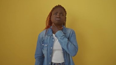 Kot pantolon giyen, grip belirtilerinden muzdarip, izole edilmiş sarı bir arka planda duran, boyun enfeksiyonu yüzünden ağrıyan boğazına dokunan Afrikalı Amerikalı bir kadın.