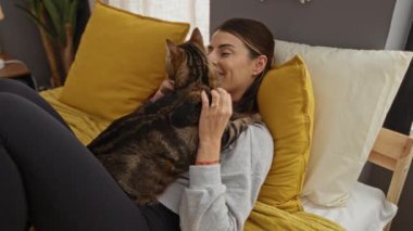 Genç bir kadın, evcil bir hayvanla sıcak ve samimi bir ev ortamını gösteren, kapalı bir yatak odasında kedisine sarılıyor..
