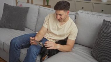 Genç İspanyol bir adam modern bir oturma odasında rahat bir şekilde kanepede tekel köpeğini okşarken akıllı bir telefon kullanıyor..
