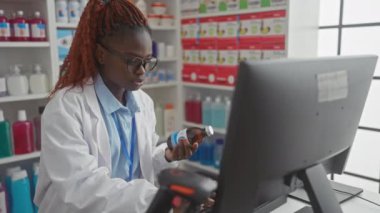 Genç bir Afrikalı eczacı iyi stoklanmış bir eczanede bilgisayar üzerinde çalışırken ilaç şişesini analiz eder..