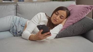 Oturma odasındaki genç bir kadın gri bir kanepede pembe bir yastıkla uzanıyor, rahat ve uykulu görünürken akıllı telefonunu kullanıyor..