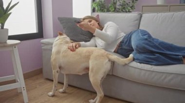 Genç bir kadın evcil köpeğiyle bir kanepede uzanıyor, rahat bir oturma odasında, uzaktan kumandayı tutuyor ve hayvanla oynuyor..