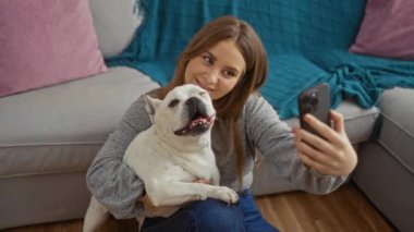 Uzun sarı saçlı genç bir kadın, rahat bir oturma odasında gri bir kanepe ve renkli yastıklarla otururken beyaz köpeğiyle selfie çeker..