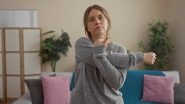 Genç sarışın bir kadın, rahat bir oturma odasında, gri bir süveterle, rahat bir kanepede canlı yastıklar ve yapraklı bitkilerle geriniyor..