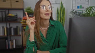 Genç sarışın kadın modern bir ofis ortamında çalışıyor. Gözlük ve yeşil gömlek takıp gülümsüyor..