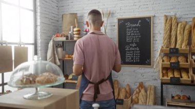 Fırında çalışan genç bir adam taze ekmek ve hamur işleriyle dolu bir kese kağıdı hazırlıyor, renkli çörekler ve ekmeklerle dolu beyaz tuğla bir duvarın önünde duruyor.