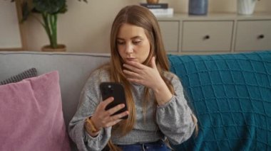 Genç sarışın, beyaz bir kadın, oturma odasında otururken akıllı telefon kullanıyor. Düşünceli bir ifadeyle..