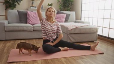 Genç bir kadın oturma odasındaki pembe bir paspasın üzerinde yoga yapıyor, yanında küçük chihuahua köpeği var, rahat iç mekanı bir kanepe ve serin bir ev egzersizi için bitkilerle tamamlıyor..