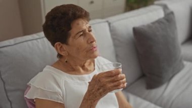Yaşlı bir kadın evinde kanepede dinleniyor, rahat bir oturma odasında su içiyor..