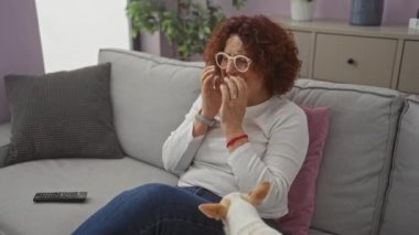 Kıvırcık saçlı ve gözlüklü yaşlı bir kadın, oturma odasında kucağında bir chihuahua otururken ellerini yüzüne koyup koltukta oturuyor..