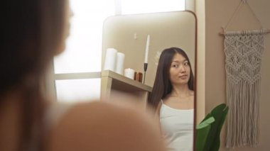 Beyaz kolsuz tişörtlü, uzun saçlı bir kadın spa aynasında kendine bakıyor. Kapalı bir ortamda güzellik ve sağlığını yansıtıyor.