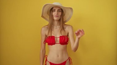 Bikini giymiş, yaz şapkalı beyaz bir kadın izole edilmiş sarı arka planda parmaklarından bakarak mutlu jestler yapıyor, gülümsüyor ve güneşte eğleniyor.