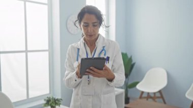 Laboratuvar önlüklü, odaklanmış bir İspanyol kadın doktor modern bir klinikte doğal ışığı olan bir tablet kullanıyor..