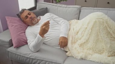 Orta yaşlı bir adam oturma odasındaki kanepede hasta gibi görünüyor ve evde yatarken termometreyle ateşini ölçüyor..