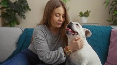 Sarışın bir kadın, rahat bir oturma odasında gülümseyen beyaz köpeğini kucaklıyor. Gri bir kanepede oturuyor. Etrafı canlı bitkilerle çevrili..