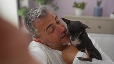 Orta yaşlı İspanyol bir adam arka planda bitkilerle sıcak bir salonda köpeğine sarılıyor..