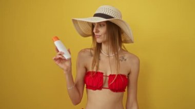 Bikinili kadın elinde güneş kremi tutarken şaşırmış ve mutlu bir yüzle, parmağını kaldırmış poz veriyor ve sarı arka plandaki ifadeyi sorguluyor.