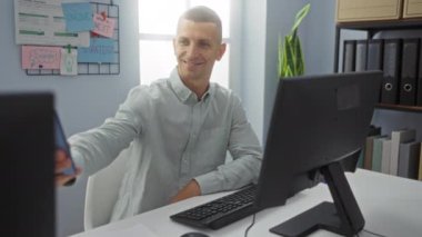 Ofisteki genç adam masadaki akıllı telefonuyla özçekim yapıyor bilgisayar vitrinleri çalışma alanı, strateji notları ve günlük kıyafetlerle.