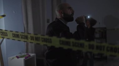 Bir adam ve bir kadın, muhtemelen dedektifler, kapalı bir suç mahallini, uyarı bandı ve el fenerleriyle, loş ışıkta inceleyin..