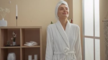 Genç ve güzel bir kadın, spa odasında bir anlık dinlenmenin keyfini çıkarıyor. Beyaz bir bornoz ve kafasında havluyla, rahat bir dekor ve doğal ışıkla..