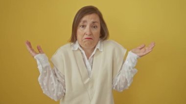 Orta yaşlı İspanyol kadın, soyutlanmış sarı arka plana karşı duruyor. İfadesiz bir ifadeyle, kollarını ve ellerini kaldırmış, şüphe ve kafa karışıklığını somutlaştırıyor.