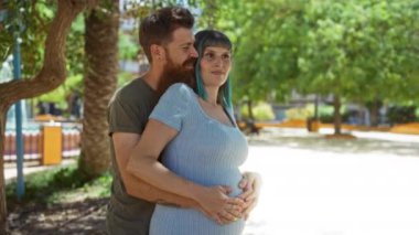 Hamile kadın ve erkek, güneşli bir şehir parkında kucaklaşıyorlar. Çevrelerinde yeşil ağaçlar ve modern şehir manzarası var.
