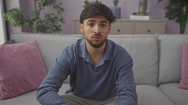 Genç, sakallı bir adam, modern bir apartman dairesinin gri koltuğunda gelişigüzel oturuyor..